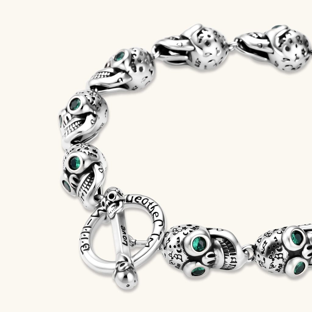 Silver stainless steel skull link bracelet with green crystal eye sockets and engraved detailing, toggle clasp closure.