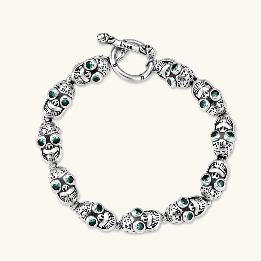 Silver stainless steel skull link bracelet with green crystal eye sockets and engraved detailing, toggle clasp closure.